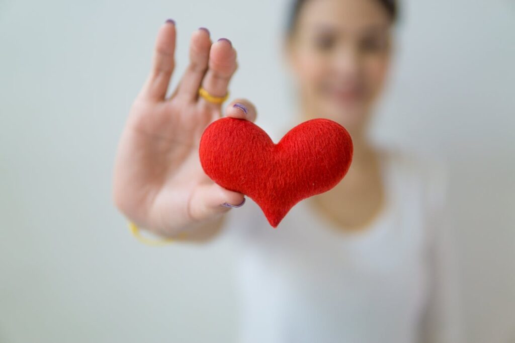 Unrecognizable blurred female standing on white background with outstretched arm while demonstrating red decorative heart in light studio in daytime