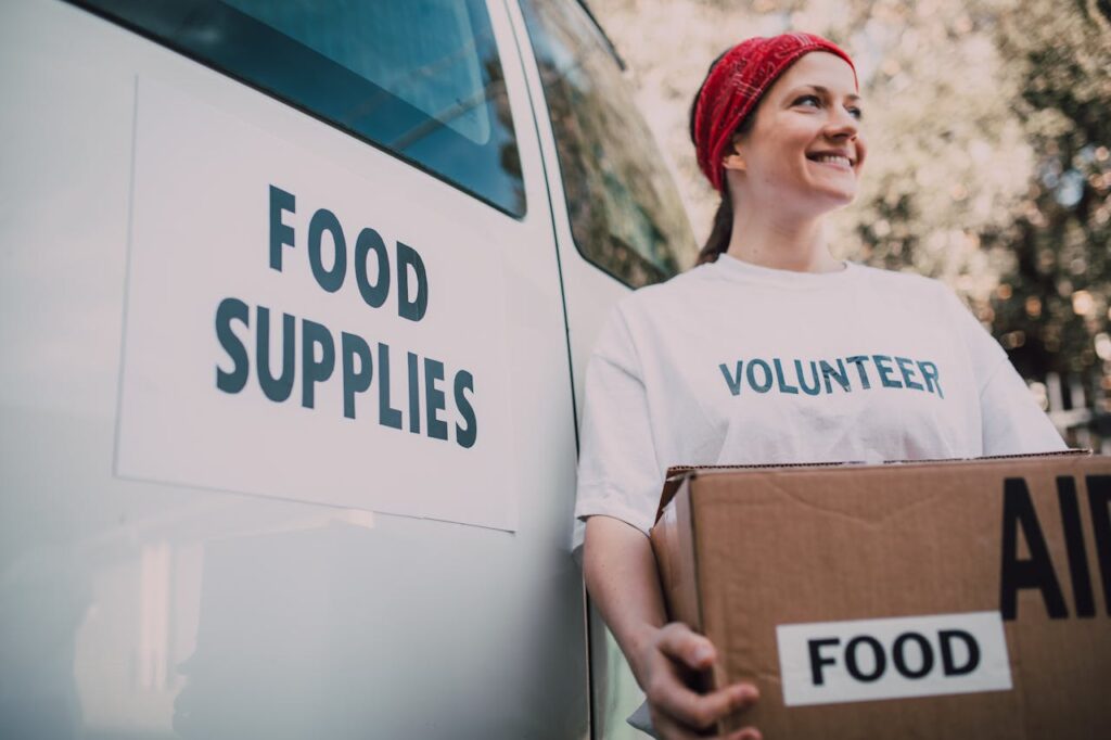 Smiling volunteer with a box of food supplies next to a delivery van.