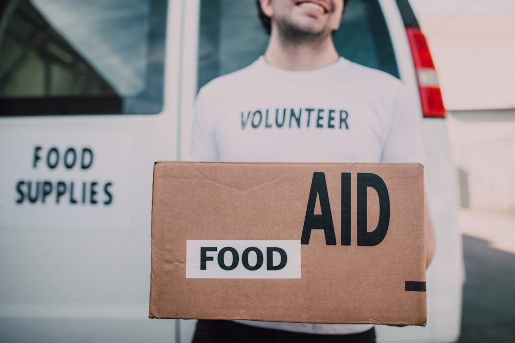 Smiling volunteer holding a box labeled 'Food Aid' outside a van with 'Food Supplies' sign.
